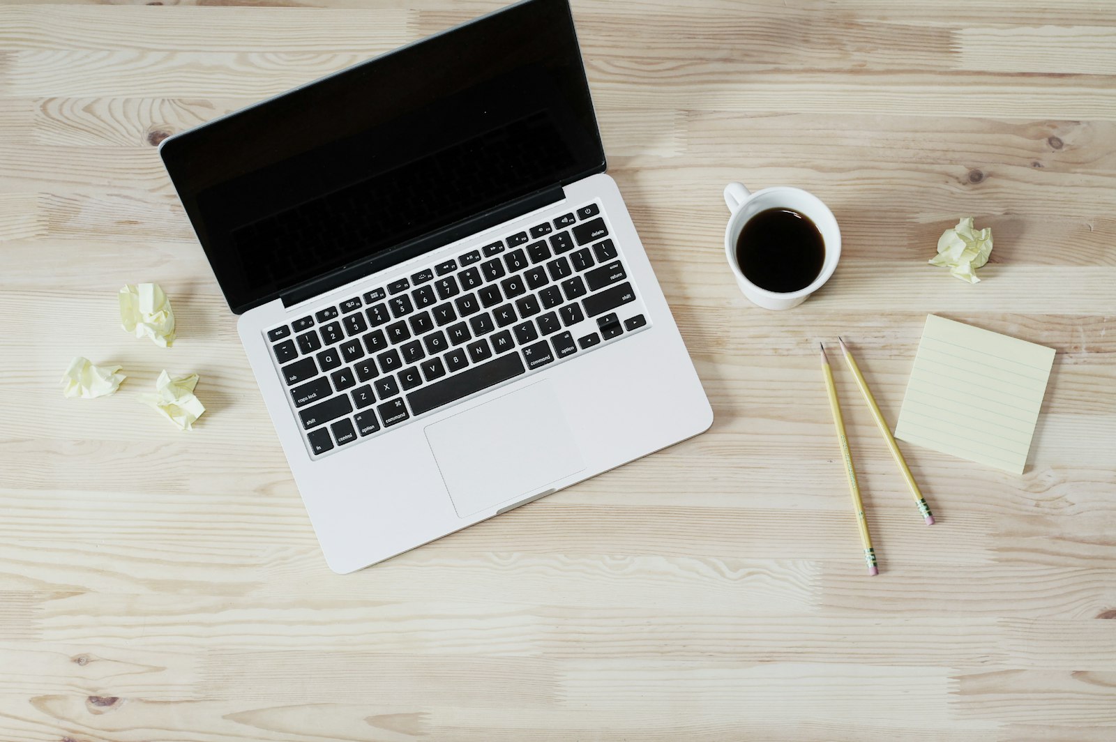 Overhead view of a MacBook on a pale wood table with coffee and pencils