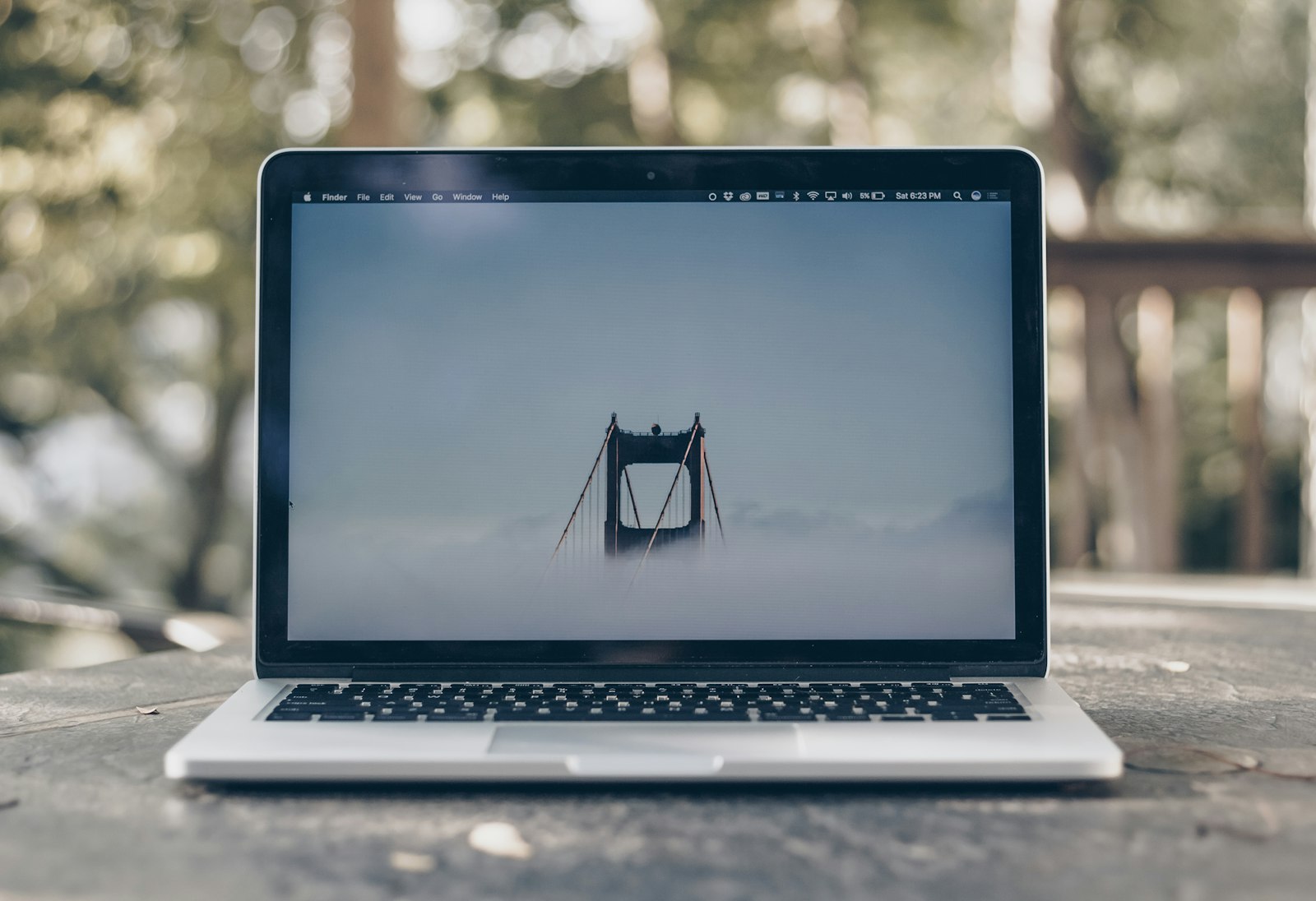 MacBook on an outdoor table showing a bridge wallpaper in macOS